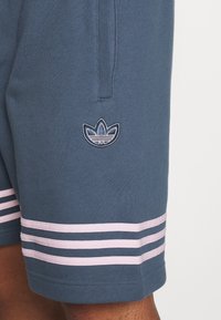 Tmavě modré sportovní šortky vyrobené z měkké tkaniny, s třemi bílými a růžovými vodorovnými pruhy na spodním okraji a vyšívaným logem.