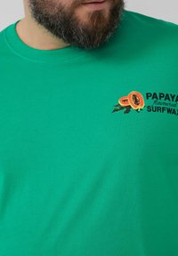 Groene katoenen t-shirt met een papaja-afbeelding en de tekst "PAPAYA smaak SURFWARE" op de voorkant. Simpel en casual ontwerp.