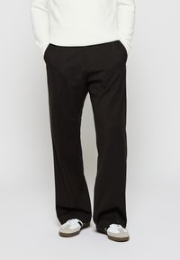 Persona melnā plašu biksēs ar rokām kabatās, baltu džemperi un baltām sporta kurpēm ar pelēkiem un melniem akcentiem, stāv uz baltas fona.