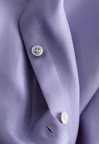 Gros plan d’un tissu de chemise lavande avec deux boutons blancs et des coutures visibles le long de la patte de boutonnage et de la boutonnière.