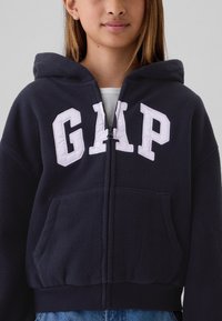 Mörkblå fleecetröja med huva, känguruficka, dragkedja och vit "GAP"-logga på bröstet; har snörning i luvan och ribbade muddar.