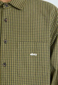 Groen en gele geruite blouse met een borstzak. Kenmerkt zich door bruine knopen en een wit "obey" logo op de zak.