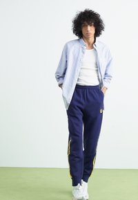 Conjunto casual com uma camisa de botão riscada em azul claro, uma t-shirt branca e calças de fato azul-escuras com riscas amarelas laterais. Ténis brancos.