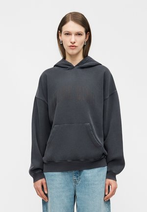 Γυναίκα που φοράει σκουρόχρωμο, υπερμέγεθες φούτερ με μπροστινή τσέπη και ξεθωριασμένο κείμενο, συνδυασμένο με ανοιχτόχρωμα μπλε τζιν, στέκεται μπροστά σε απλό λευκό φόντο.