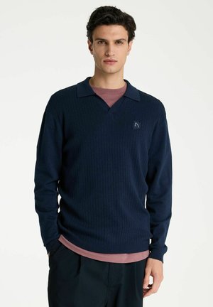 Ein Mann mit dunklen Haaren trägt einen marineblauen gerippten Pullover über einem staubrosafarbenen Hemd und steht mit einer Hand in der Tasche vor einem weißen Hintergrund.