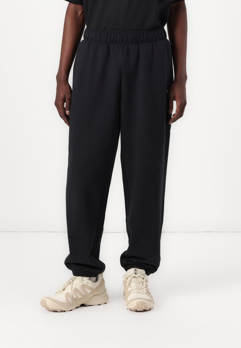 Sorte joggebukser med elastisk liv, sidelommer, kombinert med beige treningssko med teksturdetaljer og snøring.