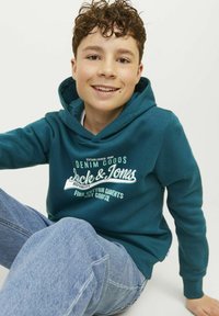 Felpa con cappuccio teal con testo grafico bianco, abbinata a jeans azzurri chiari. Tessuto morbido, vestibilità comoda, dettagli di cucitura visibili.