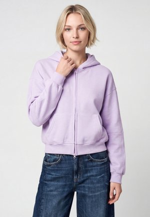 Giovane donna con capelli biondi corti che indossa una felpa con zip lilla chiaro e jeans blu su uno sfondo grigio uniforme.