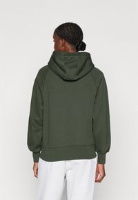 Hoodie verde închis cu guler înalt și croială relaxată. Dispune de mâneci raglan și manșete ribbate, fabricat dintr-un material moale cu o textură fină.