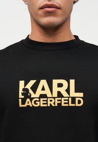 Κοντινό πλάνο ατόμου που φοράει μαύρο πουκάμισο με χρυσό κείμενο "KARL LAGERFELD" και σιλουέτα λογοτύπου στο στήθος.