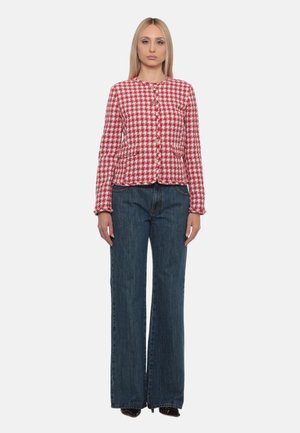 Donna con capelli biondi lisci indossa una giacca a quadri rossa e bianca e jeans a gamba larga blu scuro, in piedi davanti a uno sfondo uniforme.