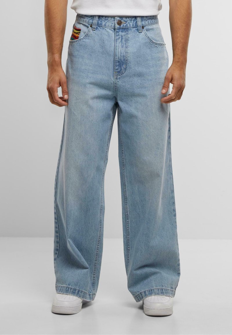 Mann trägt weit geschnittene, hellblaue Jeans mit einem kleinen, bunten Aufnäher auf der vorderen Tasche und weißen Turnschuhen.