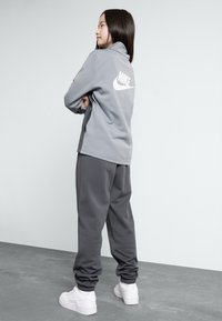 Pullover gris Nike avec logo blanc au dos, associé à un jogging gris. Tissu lisse, poignets élastiques et baskets blanches complètent le look.