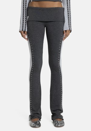 Kvinde iført mørkegrå tætsiddende bukser med sølvnitter langs siderne og matchende grå ballerina-sko, stående mod en hvid baggrund.