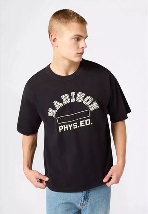 Giovane uomo che indossa una maglietta nera con la scritta "Madison Phys. Ed.", guarda di lato, regge l'orlo della maglietta, con jeans azzurri chiari su sfondo semplice.