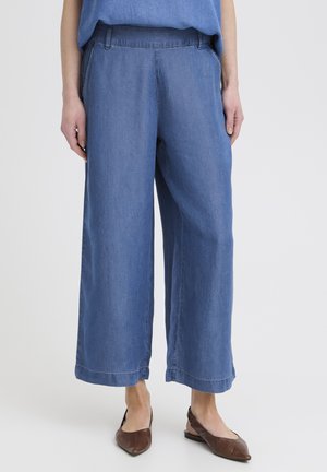 Donna con pantaloni corti di jeans blu larghi e scarpe basse marroni con punta appuntita, in piedi su sfondo bianco.