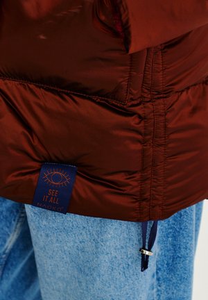Burgundská puffer bunda s hladkou, lesklou texturou. Obsahuje modrou etiketu s nápisem "VIDĚT VŠE NAOKO" a stahovací šňůrku v dolní části.