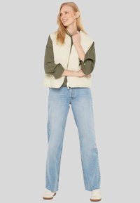 Leichter, steppverarbeiteter cremefarbener Weste über einem dunkelgrünen Hemd, kombiniert mit hellblauen, weitgeschnittenen Jeans und weißen Sneakers. Schlichtes, lässiges Design.