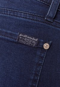 Jeans de mezclilla azul oscuro que presentan una pequeña etiqueta negra con texto blanco y un detalle de botón plateado cerca del bolsillo. Textura suave con detalle de costura.