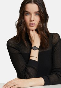 Orologio da polso nero con quadrante rotondo e accenti argentati, dotato di un bracciale in rete metallica. Indossa un top nero a maniche lunghe e a coste.