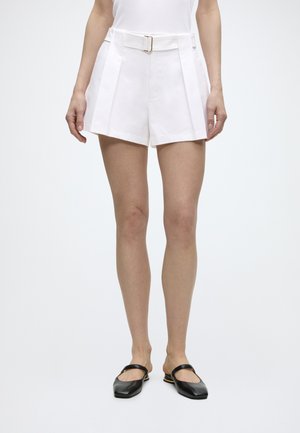 Person iført hvide bælte-pleated shorts og sorte Mary Jane-flade sko med firkantet tå, stående mod en ensfarvet lys baggrund.