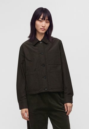 Jeune femme aux cheveux noirs et lisses portant une veste sombre boutonnée avec de grandes poches et un pantalon foncé sur un fond uni.