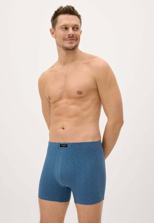 Hombre sin camiseta con el cabello corto que lleva puestos unos calzoncillos bóxer azules con estampado, de pie y mirando hacia un lado contra un fondo claro y liso.