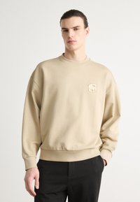Beige színű pulóver kerek nyakkal, laza fazonnal és bordás mandzsettákkal. Diszkrét, dombornyomott logó található a mellkasán. Fekete nadrággal párosítva.