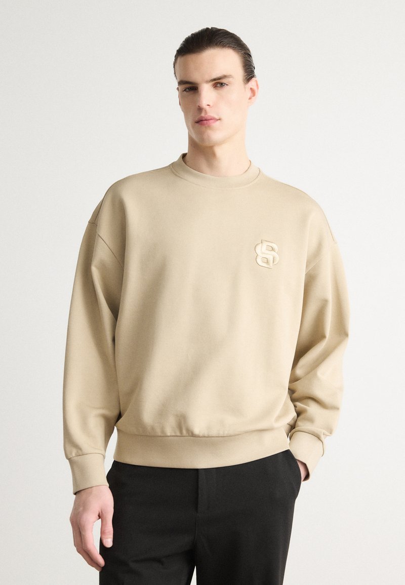 Beige színű pulóver kerek nyakkal, laza fazonnal és bordás mandzsettákkal. Diszkrét, dombornyomott logó található a mellkasán. Fekete nadrággal párosítva.