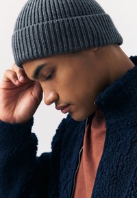 Siva rebrasta pamučna kapa na glavi modela, uparena s tamnoplavljom teksturiranom jaknom na patentni zatvarač preko majice boje hrđe. Prikaz izbliza u profilu.