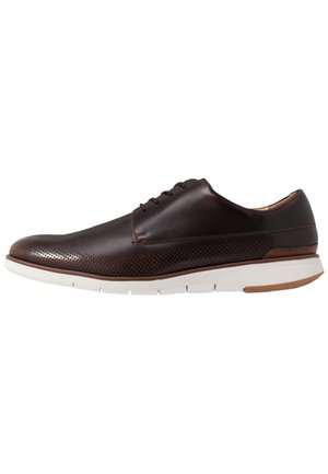 Chaussure habillée pour homme en cuir marron avec détails perforés, semelle en caoutchouc blanche et fermeture à lacets, vue de profil sur fond blanc.