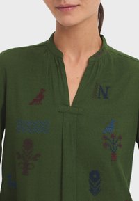 Chemisier vert avec un col montant, orné de motifs brodés d'animaux et de plantes en bordeaux et bleu.