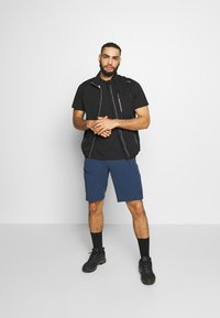 Chaleco negro con cremallera y detalles gris, camiseta negra de manga corta, pantalones cortos azules, calcetines negros y calzado negro. El modelo está de pie contra un fondo blanco.