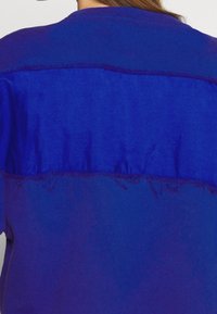 Vista trasera de una persona que lleva una sudadera azul con una franja horizontal más oscura en la parte superior de la espalda y bordes desgastados.