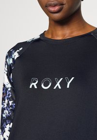 Mustapohjainen urheilupaita, jossa on värikkäät kukkakuosiset raglan-hihat. Rinnassa vaaleansinisellä "ROXY"-logo. Silkkinen ja tiukka muotoilu.