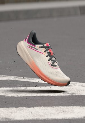 Zapatilla de running con suela degradada blanca y rosa y cordones negros, suspendida sobre una carretera de asfalto gris con rayas blancas.