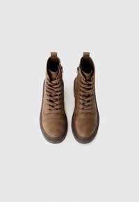 Une paire de bottes en cuir marron à lacets avec des zippers sur le côté et de épaisses semelles foncées, vues de dessus sur un fond blanc.