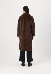 Cappotto in faux fur marrone con colletto, maniche lunghe e vestibilità ampia, abbinato a pantaloni neri a gamba larga e scarpe nere.