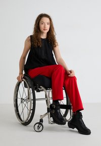 Black sleeveless top with button details, paired with red trousers. The person sits in a wheelchair wearing black ankle boots with buckles.