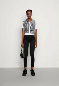Colete xadrez cortado em preto e branco sobre uma camisa branca, combinado com leggings pretas e ténis; a segurar uma pequena mala preta.