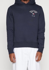 Felpa con cappuccio blu navy con logo frontale, polsini e orlo a coste, materiale in morbido cotone, con un piccolo dettaglio rosso, bianco e blu.