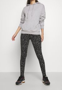 Šedá mikina s kapucí a přední kapsou, kombinovaná s černými legínami se vzory bílých abstraktních linií. Černé a vícebarevné sportovní boty.