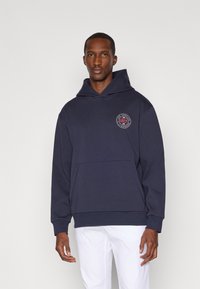 Mørkeblå hettegenser laget av mykt stoff, med en frontlomme og en sirkulær logo på venstre bryst.