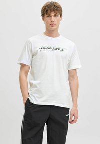 Balts īsām piedurknēm t-krekls ar melnu drukātu tekstu "JACK&JONES" un papildu grafiku. Saskaņots ar melnām biksēm, kurām ir baltas sānu svītras.