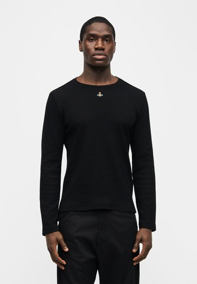 Νέος άνδρας που φοράει στενή μαύρη μακρυμάνικη μπλούζα με ένα μικρό πολύχρωμο έμβλημα κεντραρισμένο κοντά στη λαιμόκοψη, στέκεται μπροστά σε απλό φωτεινό φόντο.