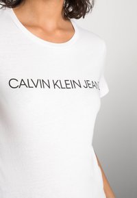 Bílý bavlněný tričko s kulatým výstřihem a krátkými rukávy, na přední straně s potiskem "CALVIN KLEIN JEANS" v tučném černém písmu.