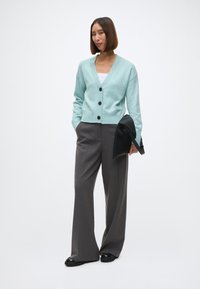 Ένα ανοιχτό τουρκουάζ πουλόβερ με V-λαιμό, ραμμένες μανσέτες και τρία μαύρα κουμπιά, συνδυασμένο με φαρδιά γκρι παντελόνια και μια μαύρη τσάντα-φάκελο.