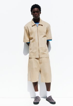 Mand iført beige kortærmet lynlåstrøje og matchende knælange shorts, med en stribet skjorte nedenunder, hvide sokker og grå sneakers, stående.