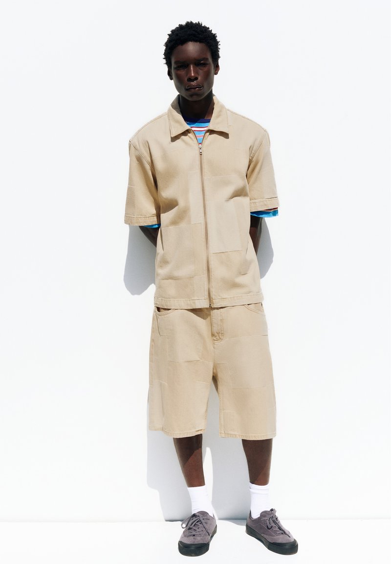 Homme portant une veste beige à manches courtes avec fermeture éclair et un short assorti longueur genoux, chemise rayée en dessous, chaussettes blanches et baskets grises, debout.