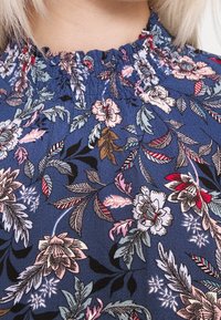 Tissu bleu avec un motif floral comportant des fleurs roses, rouges et vertes. Encolure smockée et surface texturée.